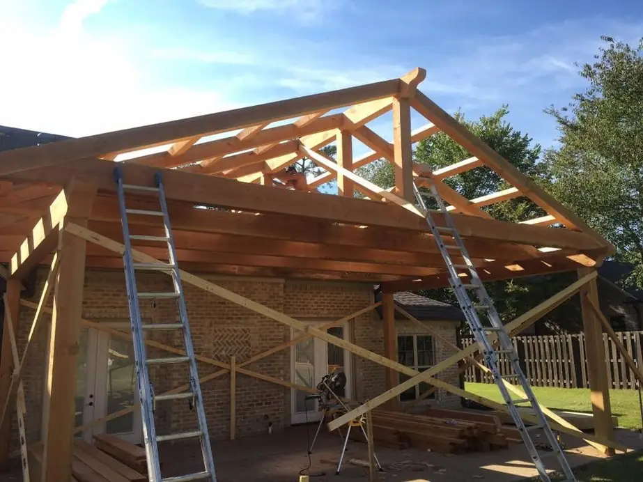 Timber porch roof framing going up against a brick house, ladders set against the beams
