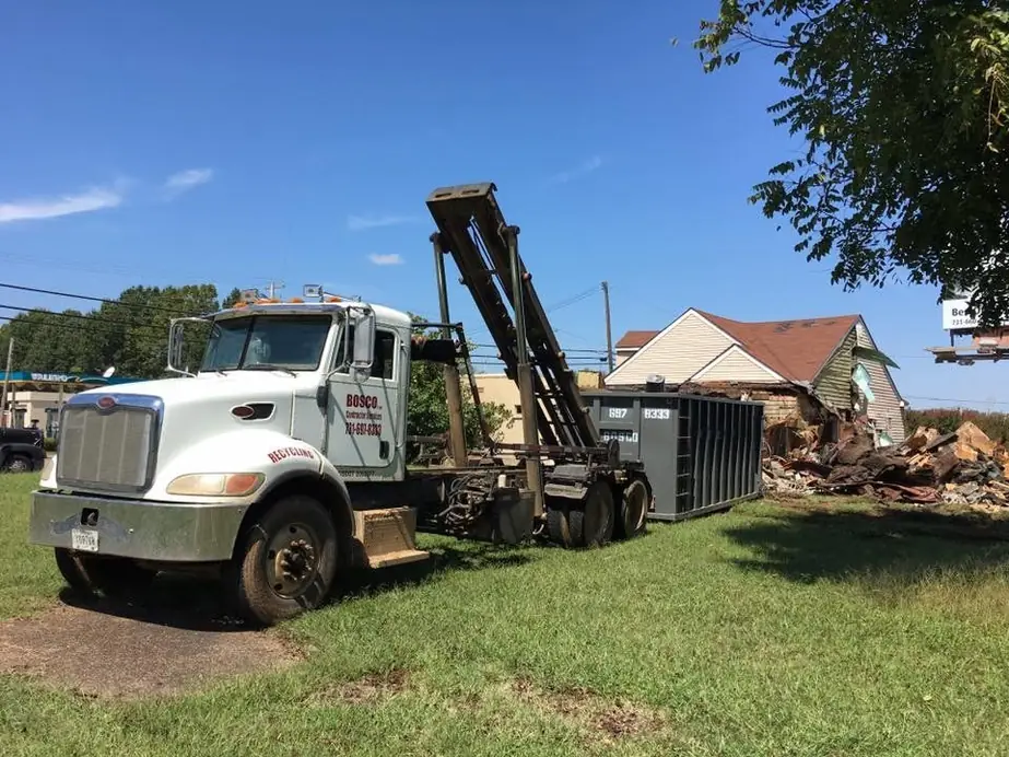 A Bosco roll-off truck with a container on the hook beside the debris of a torn-down house
