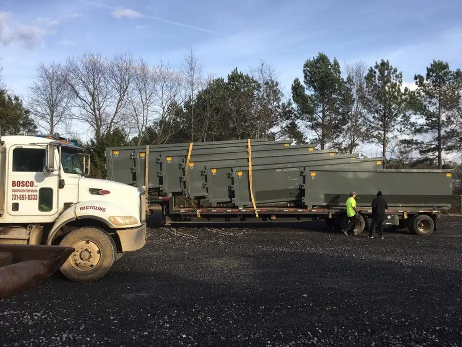 A Bosco truck delivering a flatbed load of grey containers while two crew members in hi-vis secure it