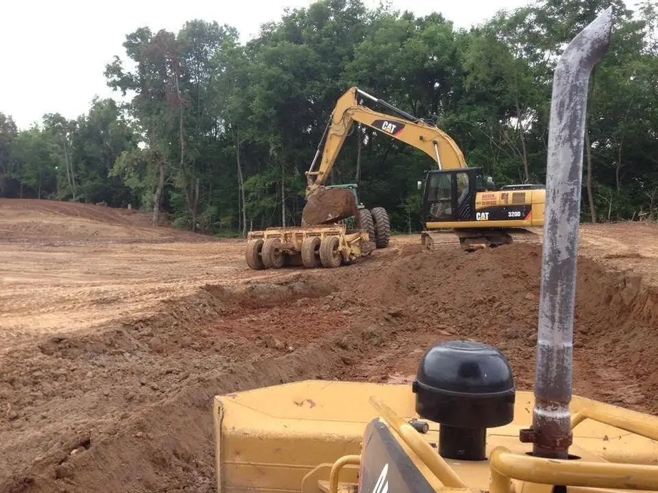 A CAT 320D excavator with a BOSCO decal and a pull scraper on a freshly graded pad, seen from the dozer seat