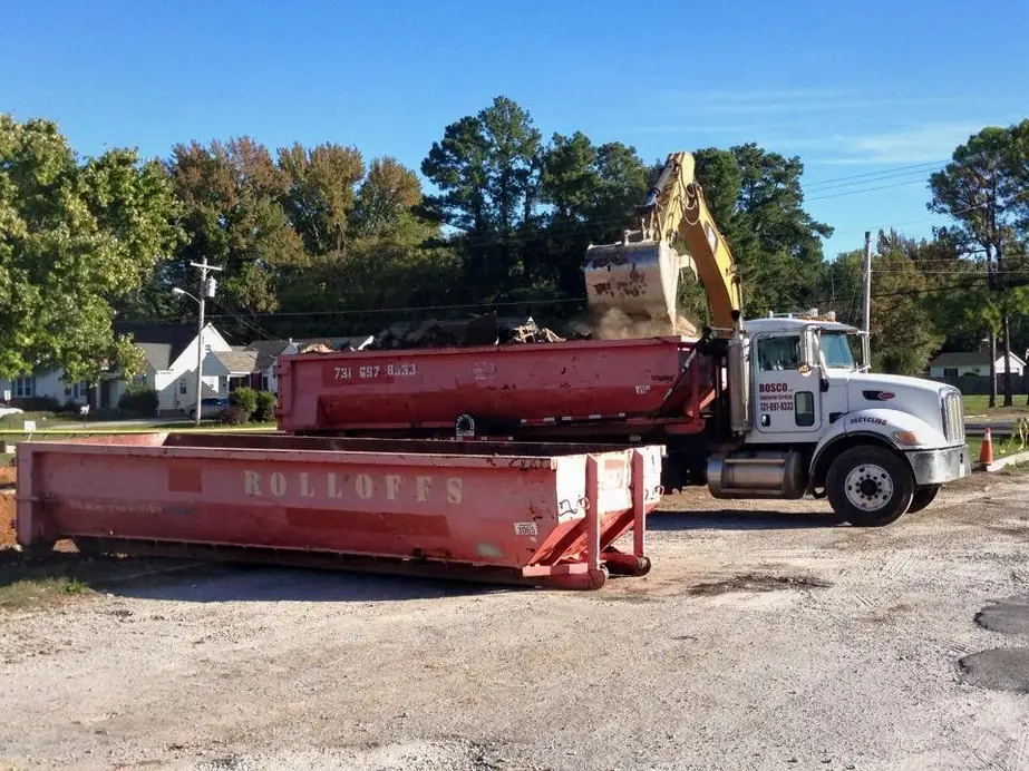Two red ROLLOFFS containers on a gravel lot with a Bosco roll-off truck and an excavator loading behind