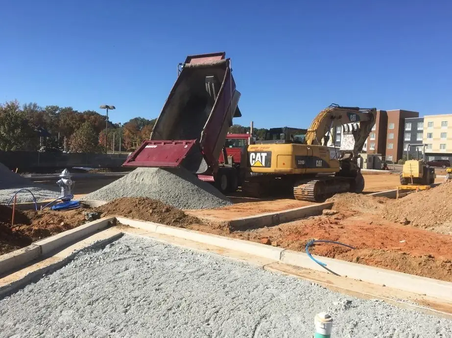 A maroon dump truck tipping gravel beside a CAT excavator on a commercial pad, curbing in the foreground