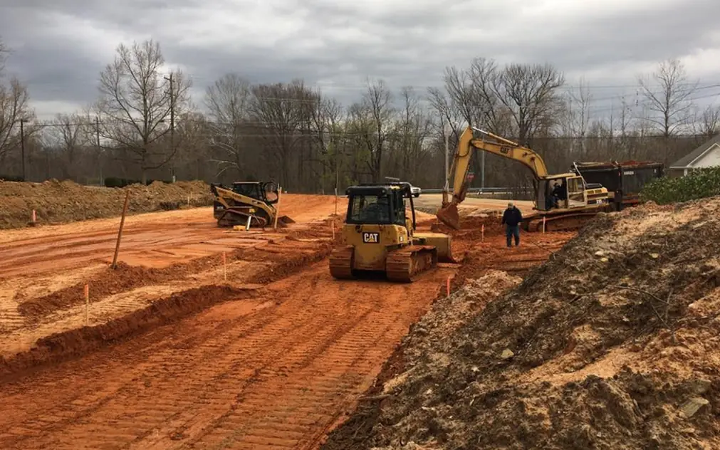 A CAT dozer, a skid steer and an excavator grading a red-clay site under a grey sky, survey stakes set in rows